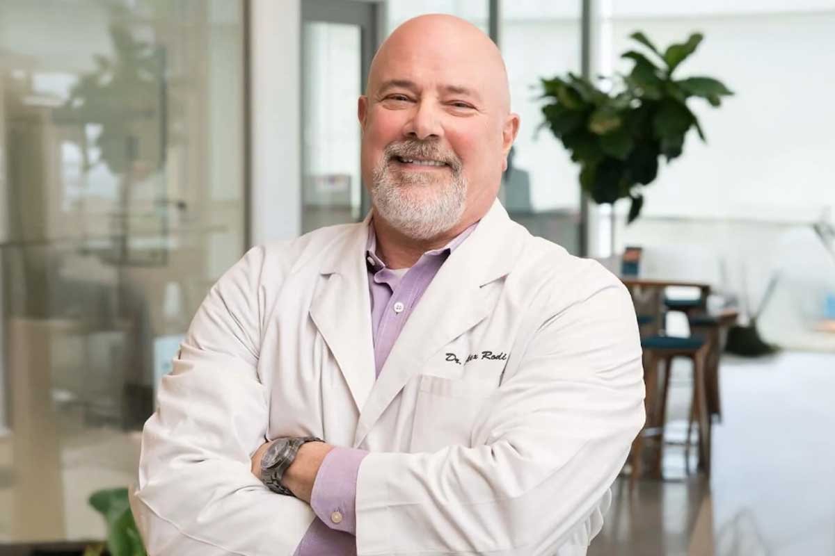 Dr. Rodi wearing a white lab coat standing with arms crossed and smiling.
