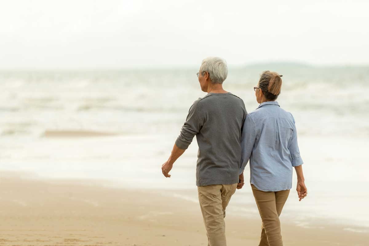 Two older adults walk hand in hand along a sandy beach, facing the ocean. The sky is cloudy, and both are dressed in casual clothes.
